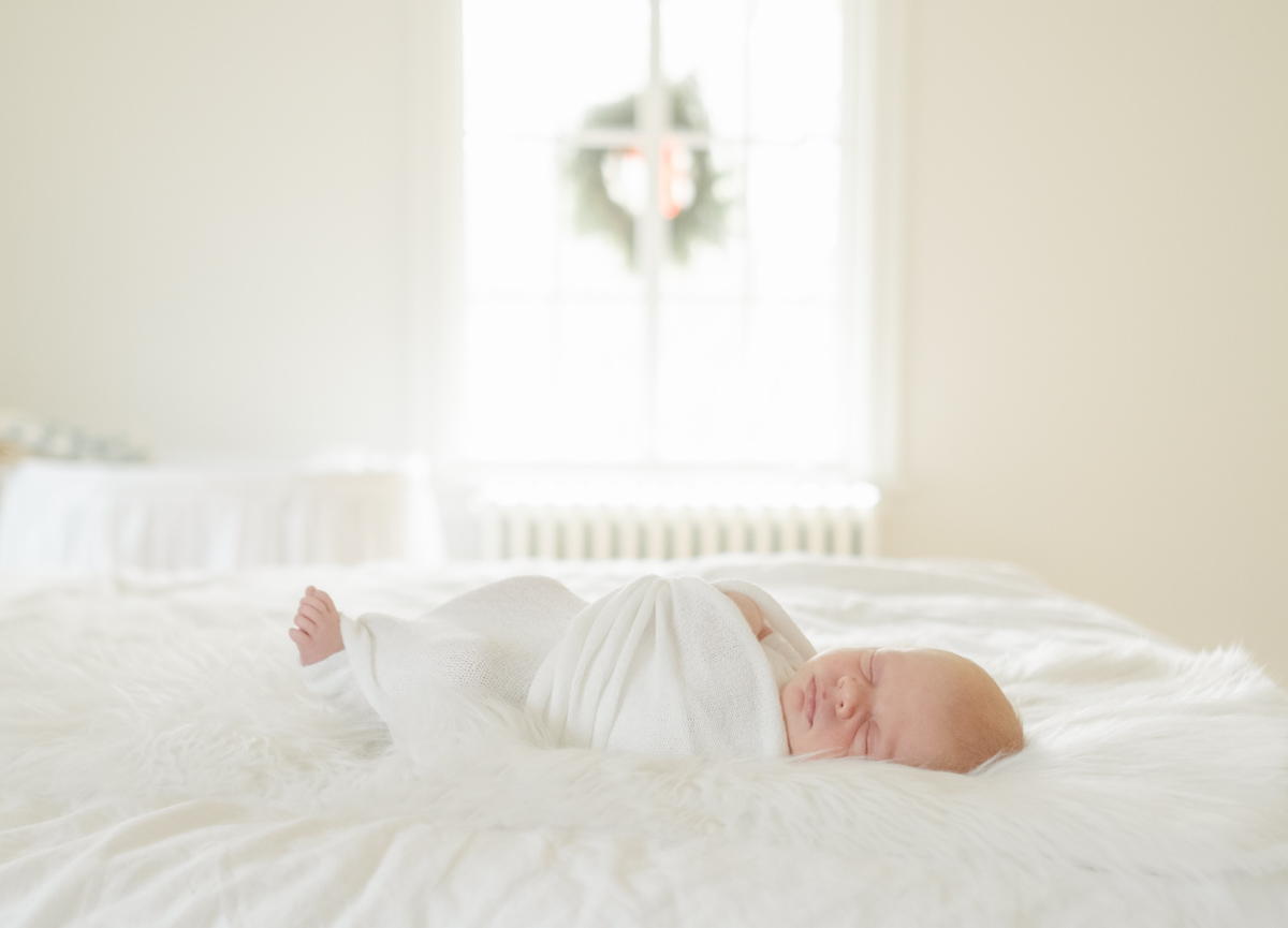 a baby wrapped in a white blanket resting on a bed a christmas wreath displays in the window behind him