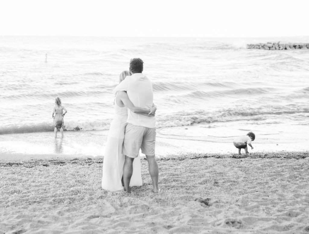 a black and white image of a mom and dad hugging while overseeing their children play on the beach in Evanston