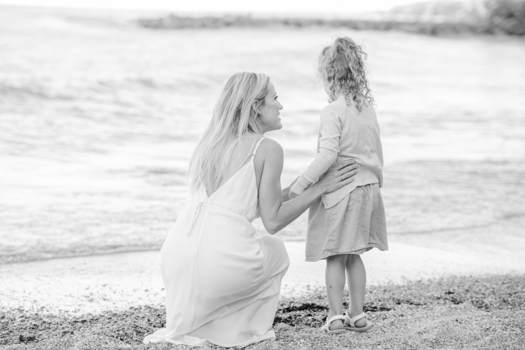 a black and white portrait of a mom crouching beside her young daughter on the beach