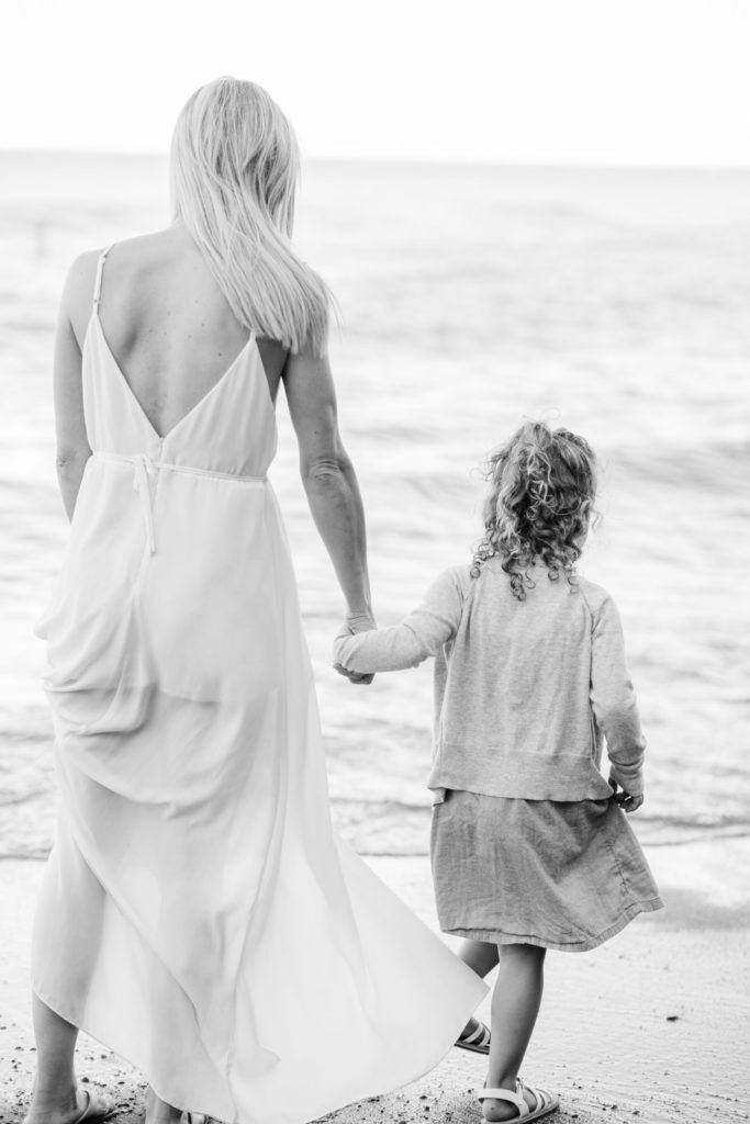 a black and white portrait of a mom and her young daughter holding hands walking down the beach