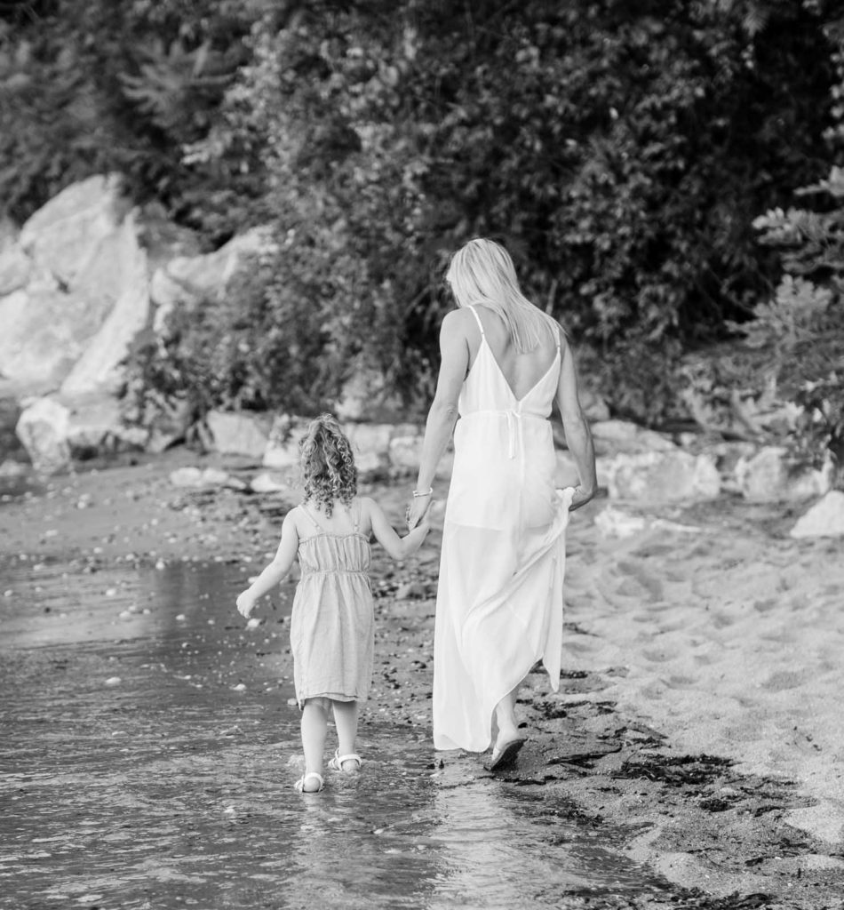 a black and white portrait of a mom and her young daughter holding hands walking down the beach
